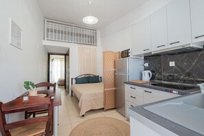 A compact hotel room with a kitchenette, a bed with a headboard, a dining area with a table and chairs, and a small balcony.