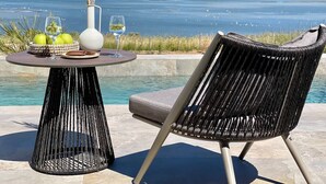 Une chaise et une table en osier, placées au bord d’une piscine, avec vue sur la mer et les montagnes.