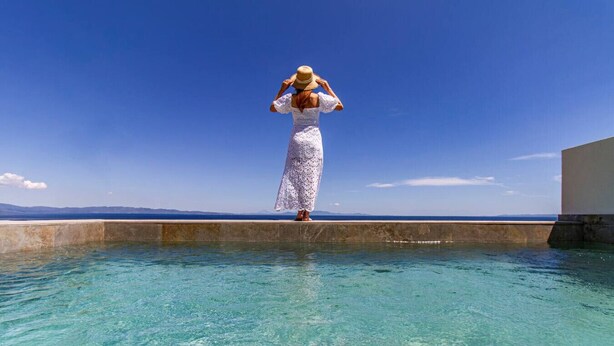 Une personne se tient près d’une piscine, d’où l’on a une vue dégagée sur la mer et les montagnes lointaines.