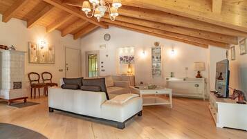 A spacious living room with a high wooden ceiling, a white sofa, a glass coffee table, and a television on a white cabinet.