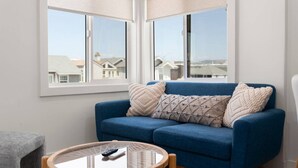Una stanza con un divano blu, due cuscini decorati, un tavolino rotondo con un telecomando e vista su case attraverso ampie finestre con tende.