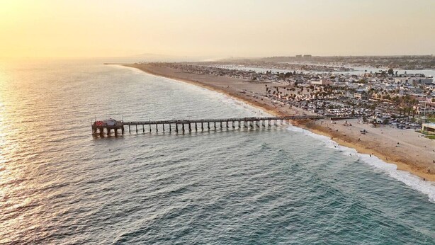 Un molo che si protende sull'oceano, con una spiaggia e sullo sfondo un paesaggio urbano.