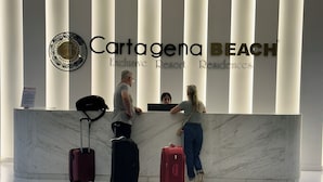 A reception desk with a logo, a man and a woman standing, and luggage. - The Suite Thirteen is a modern, minimalist apartment suite for short-term stays (Cartagena de Indias)