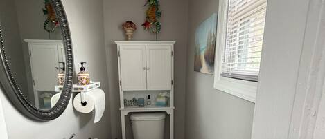 Une petite salle de bain avec un lavabo, des toilettes et un meuble de rangement blancs, un miroir et une fenêtre avec des stores.