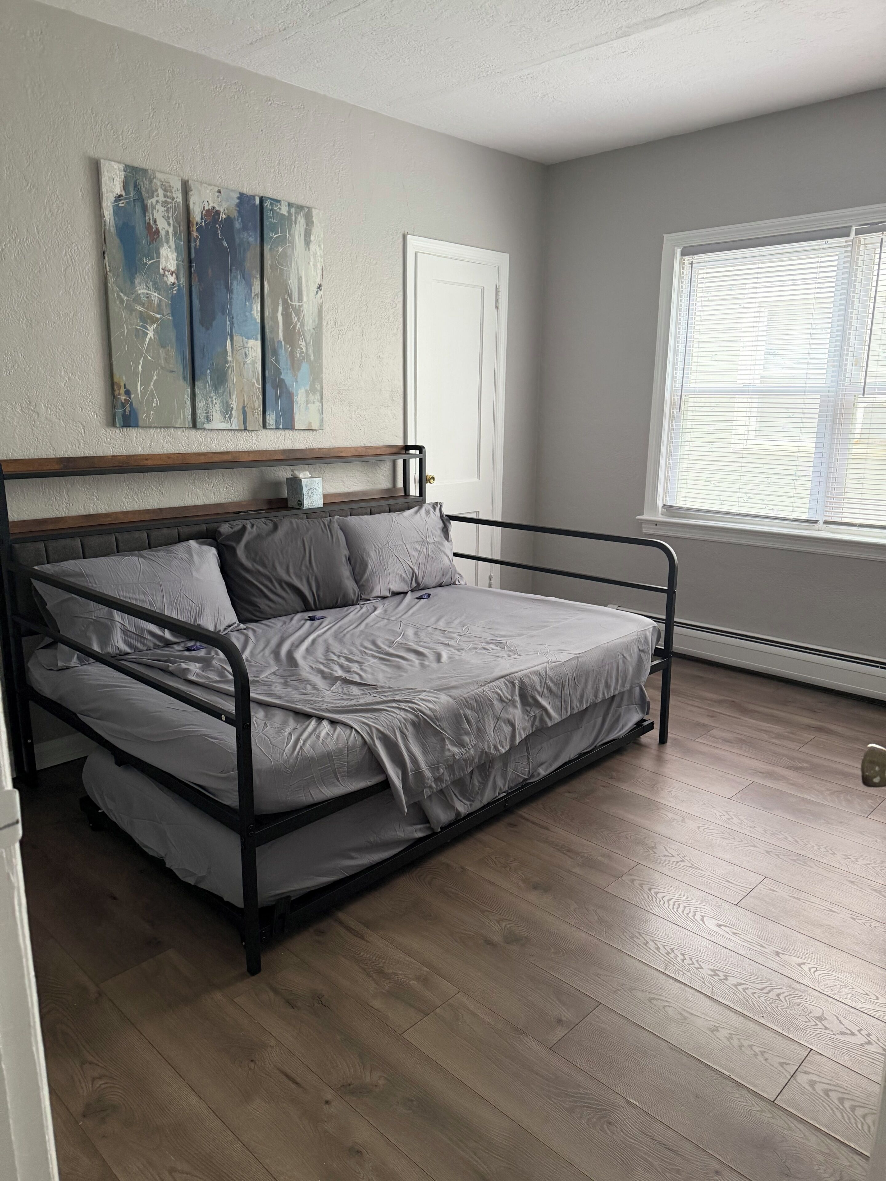 Una cama individual con estructura metálica, cabecera de madera y colchón gris en una habitación con suelo de madera y una ventana con persianas.