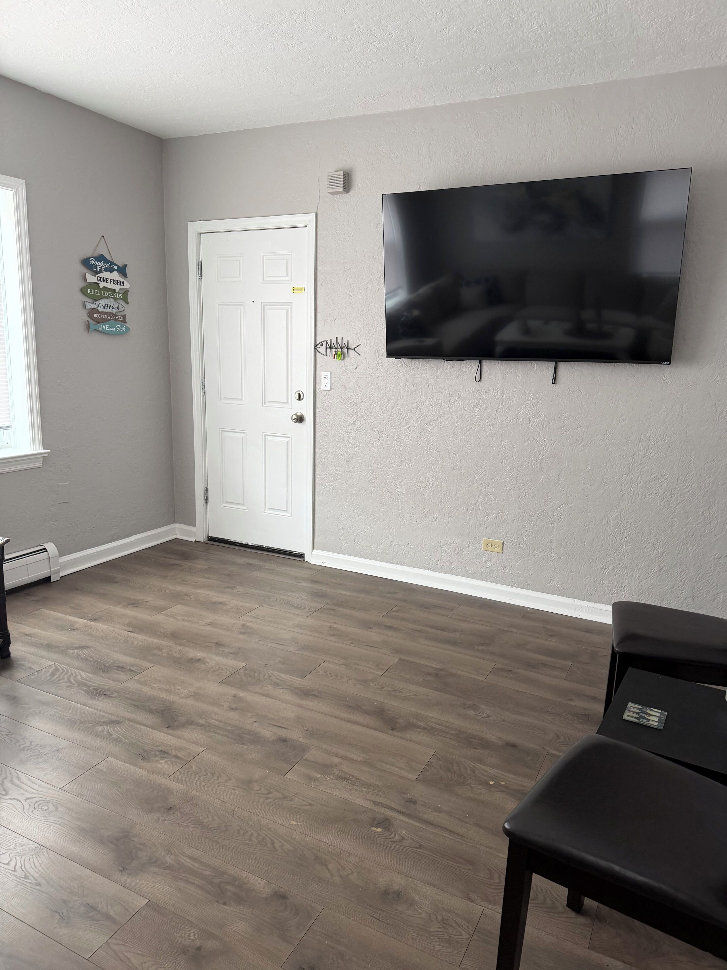 Una habitación con televisión de pantalla plana en la pared, una puerta blanca y piso de madera.