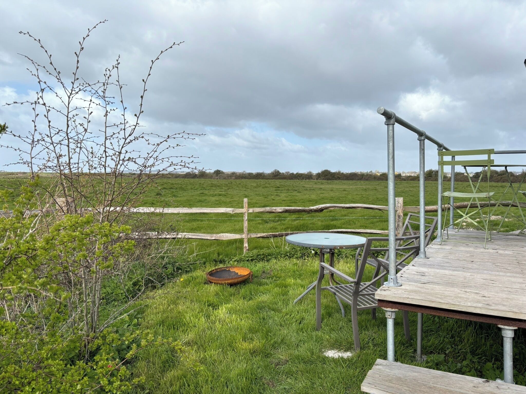 A rustic outdoor seating area with a metal table and chairs, surrounded by a grassy field and a wooden deck.