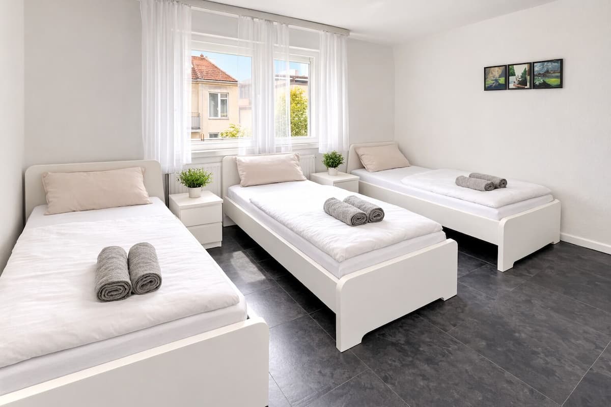 A modern hotel room with three beds, a large window, a small plant, and a wall-mounted picture.