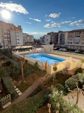 En swimmingpool omgiven av grönska, med bostadshus i bakgrunden.