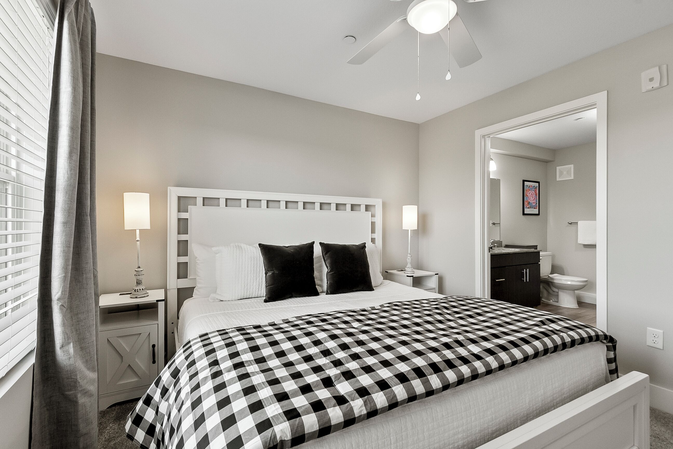 A bedroom with a checkered bedspread, a white headboard, bedside lamps, a ceiling fan, and a view into the bathroom.