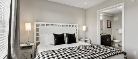 A bedroom with a checkered bedspread, a white headboard, bedside lamps, a ceiling fan, and a view into the bathroom.