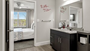 Une chambre à coucher avec un lit, un ventilateur de plafond, une fenêtre avec des rideaux, une salle de bain avec un lavabo, un miroir et un porte-serviettes.