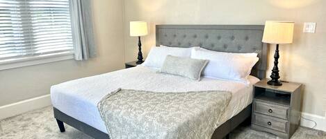 A bedroom with a bed, two bedside tables, a nightstand, a window with blinds, and a carpeted floor.