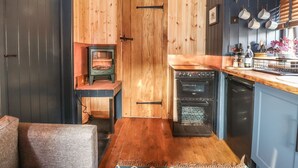 A cozy kitchen with wooden walls, a stove, oven, and a small seating area with a rug. - Dunfell Shepherd's Hut (Appleby-in-Westmorland)