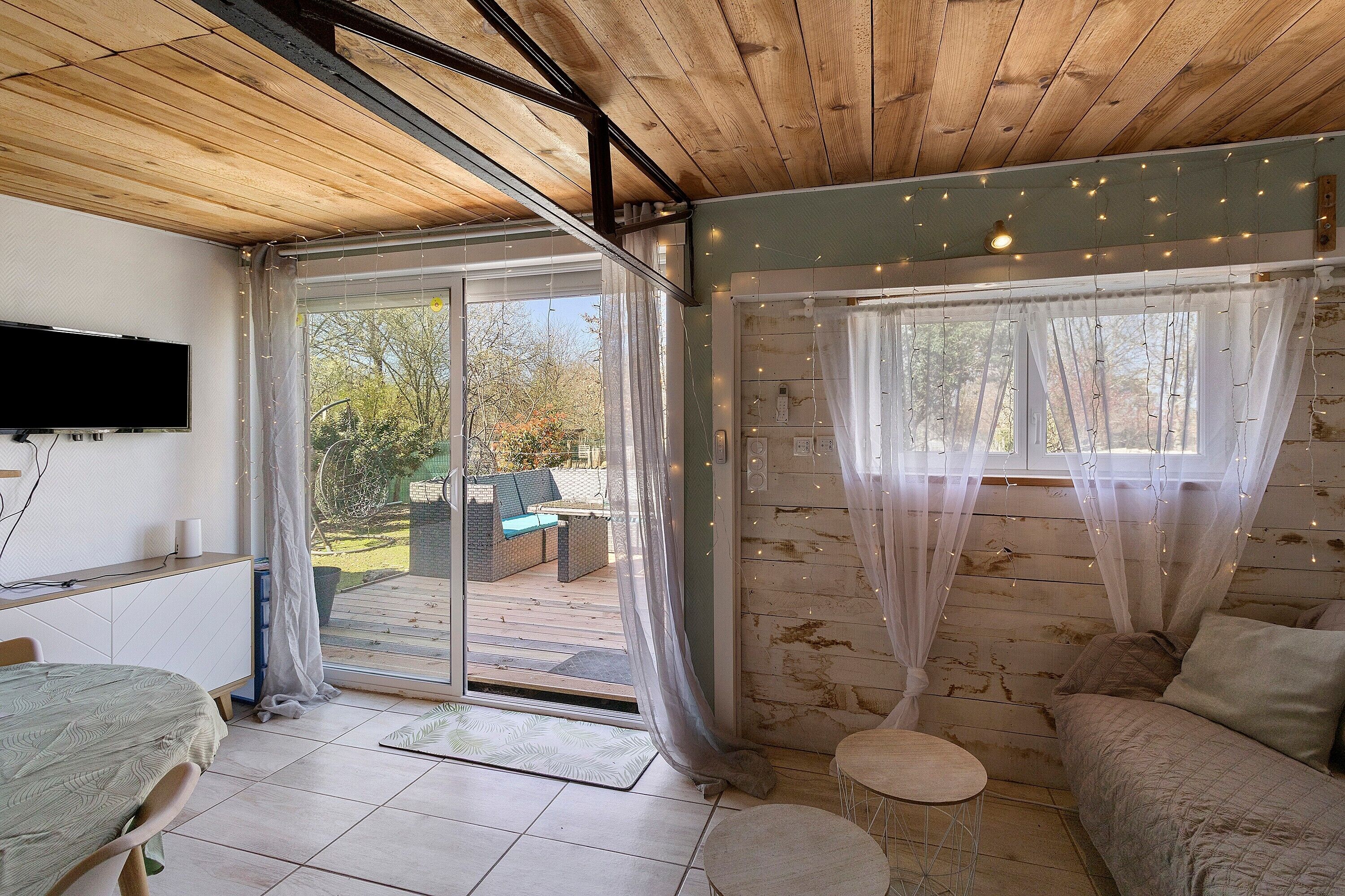 A room with a bed, a sofa, a TV, and a sliding glass door leading to an outdoor area with a patio and greenery.