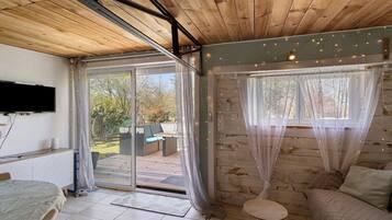 A room with a bed, a sofa, a TV, and a sliding glass door leading to an outdoor area with a patio and greenery.