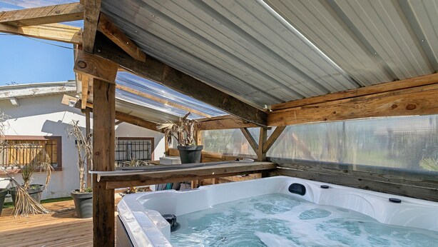 Un jacuzzi bajo una pérgola de madera con techo metálico, rodeado de plantas en macetas y una terraza de madera.
