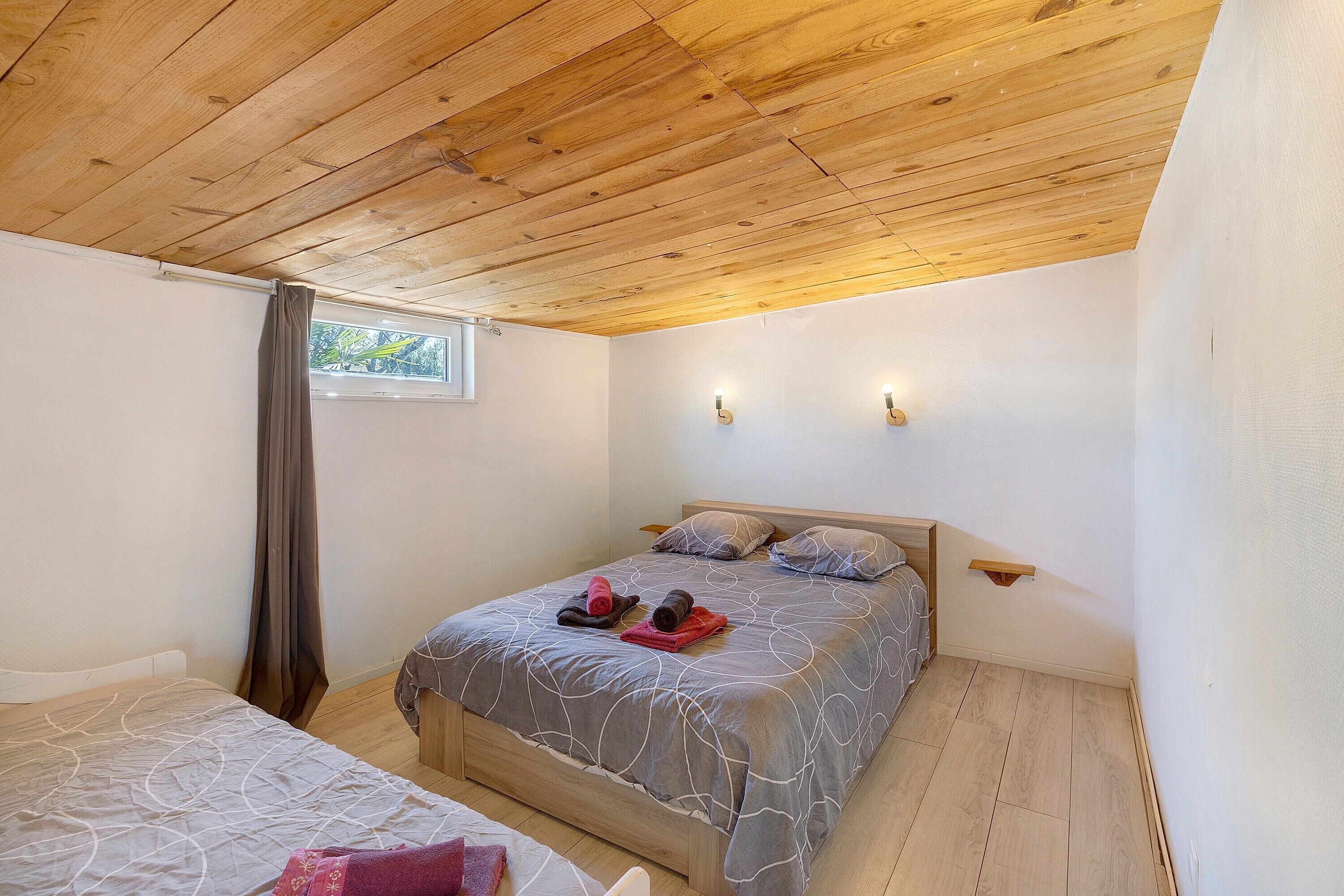Un dormitorio con techo de madera, una cama con colcha gris estampada, dos lámparas de pared y una ventana con cortina.