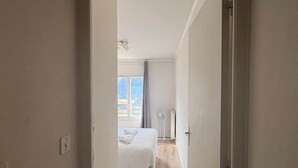 Ein schmaler Flur, der in ein Schlafzimmer mit Bett, Standventilator und einem mit Vorhängen versehenen Fenster führt.