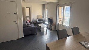 Ein modernes Wohnzimmer mit Sofa, Couchtisch und Esstisch mit Stühlen. Ein Fernseher und ein Fenster mit Vorhängen sind ebenfalls vorhanden.