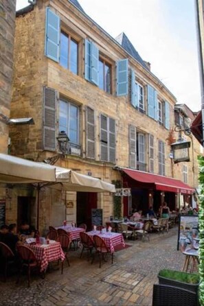 Un edificio in pietra con diverse finestre e persiane, area esterna con tavoli e sedie, e una strada acciottolata.