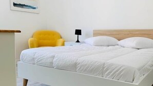 Een wit bed met een houten hoofdbord, een gele fauteuil, een nachtkastje met een lamp en een schilderij aan de muur.