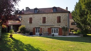 A stone building with a central entrance, a small patio with a table and chairs, and a well-maintained lawn. - L'écrin de verdure*Vue imprenable*Piscine (Cénac-et-Saint-Julien)