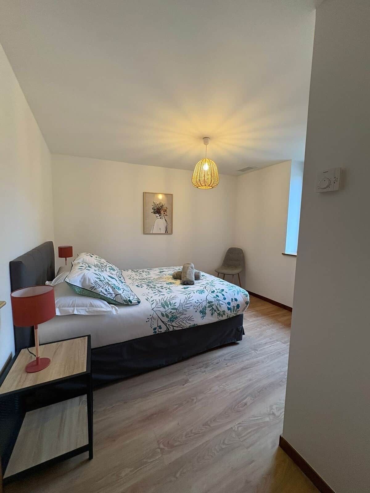 Ein Schlafzimmer mit Bett, Nachttisch mit Lampe, Stuhl, Pendelleuchte und Wandthermostat.