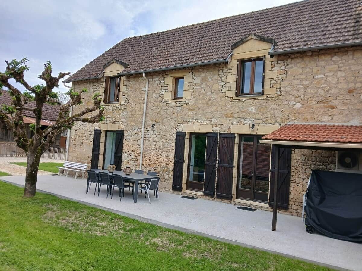 A stone building with a tiled roof, a patio with a table and chairs, and a covered area.