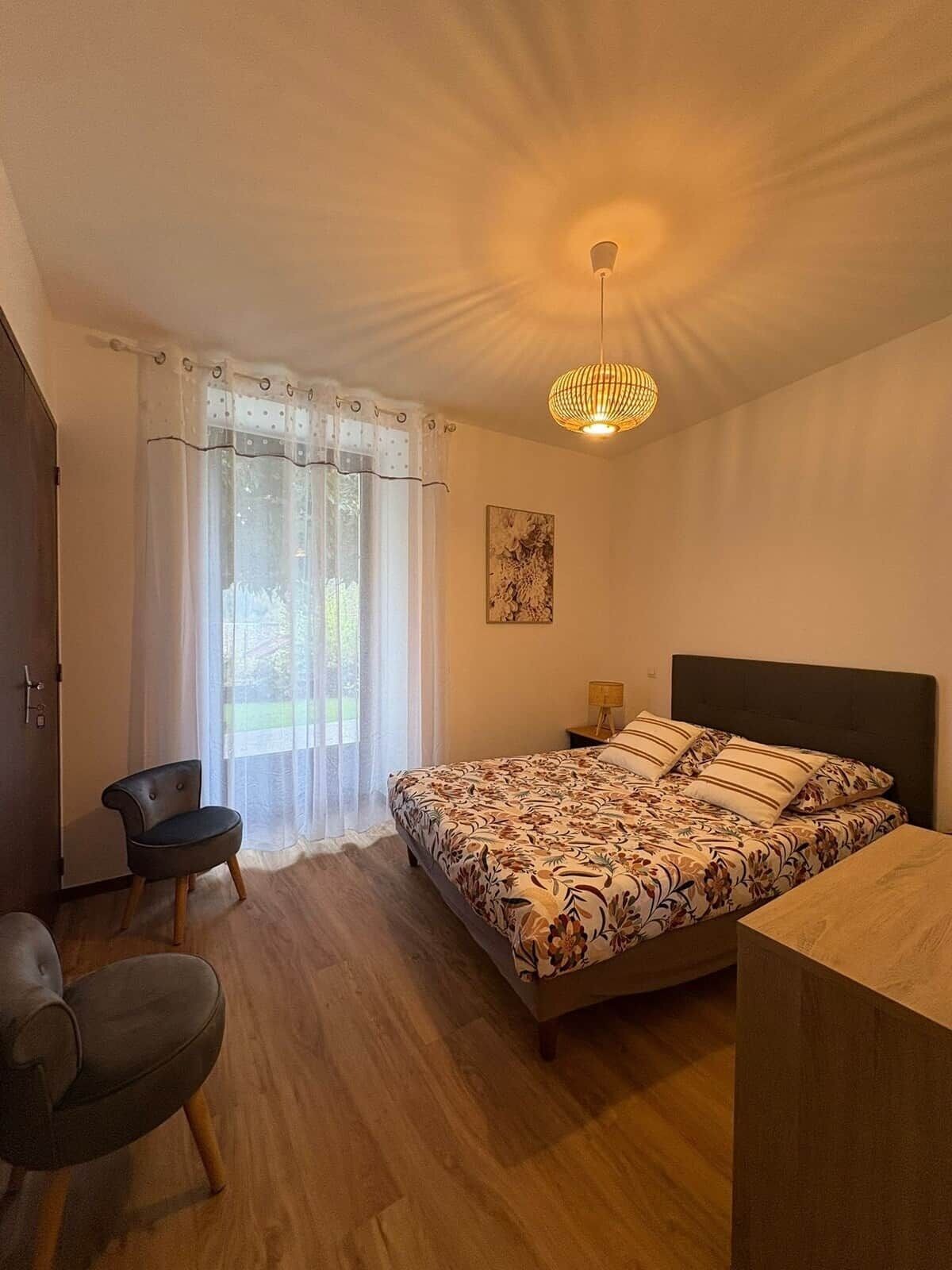 A bedroom with a bed, a chair, a bedside table, and a hanging light fixture.