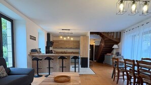 A modern kitchen with a central island, a dining area with wooden chairs, a sofa, and a staircase leading to an upper level. - Gîte des Milandes * Vue imprenable sur le Château (Castelnaud-la-Chapelle)