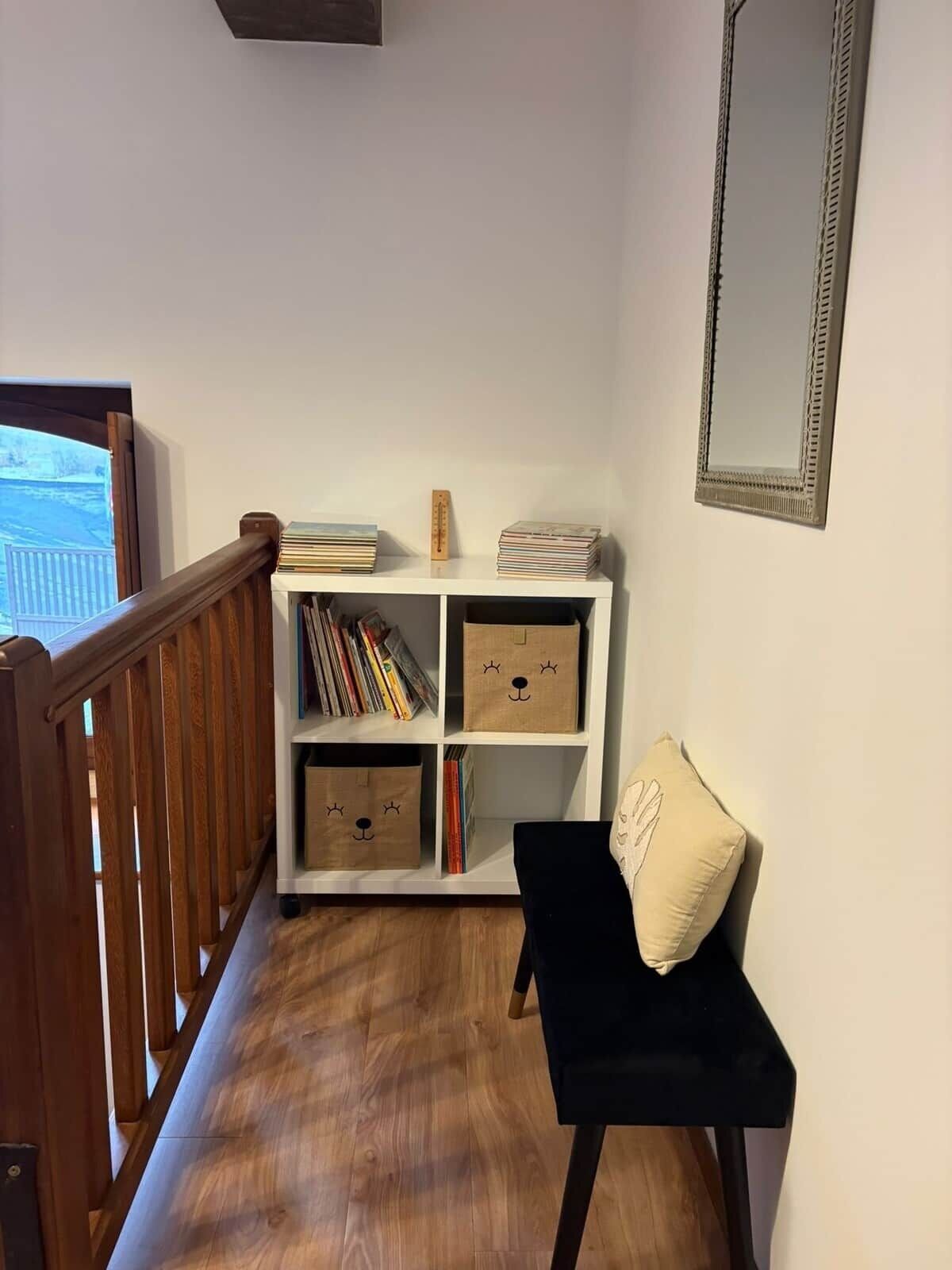 Een trap met een houten leuning, een witte boekenkast met boeken en decoratieve objecten, een zwarte stoel met kussen, en een spiegel aan de muur.