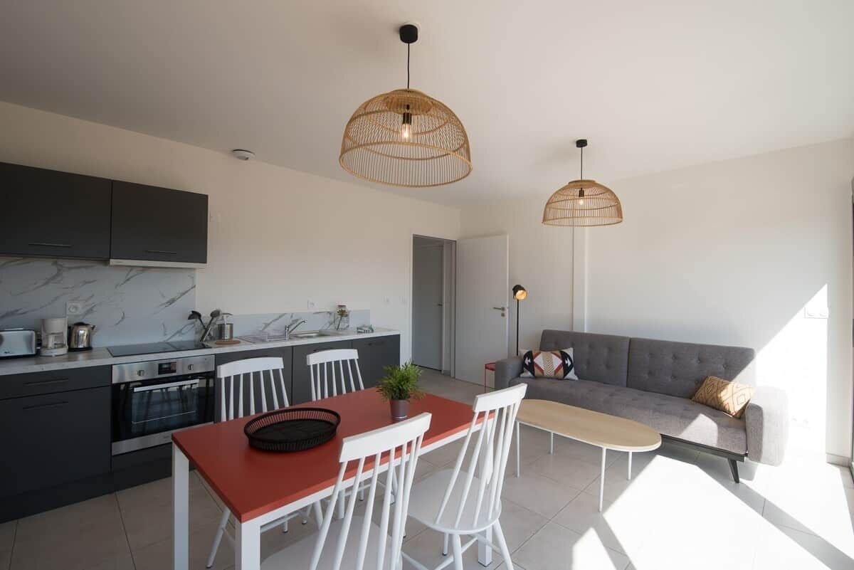 Una cocina moderna con armarios oscuros, encimera de mármol y comedor con mesa roja y sillas blancas.