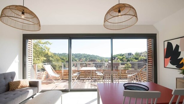 A modern living room with a sofa, a dining table, and chairs, a balcony with a view of the hills, and two hanging wicker lamps. - Parenthèse Grand Air* Centre historique* parking lot (Sarlat-la-Canéda)