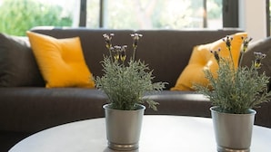 Uma sala de estar moderna com uma mesa redonda, duas plantas em vasos, um sofá marrom com almofadas amarelas e uma janela com persianas.