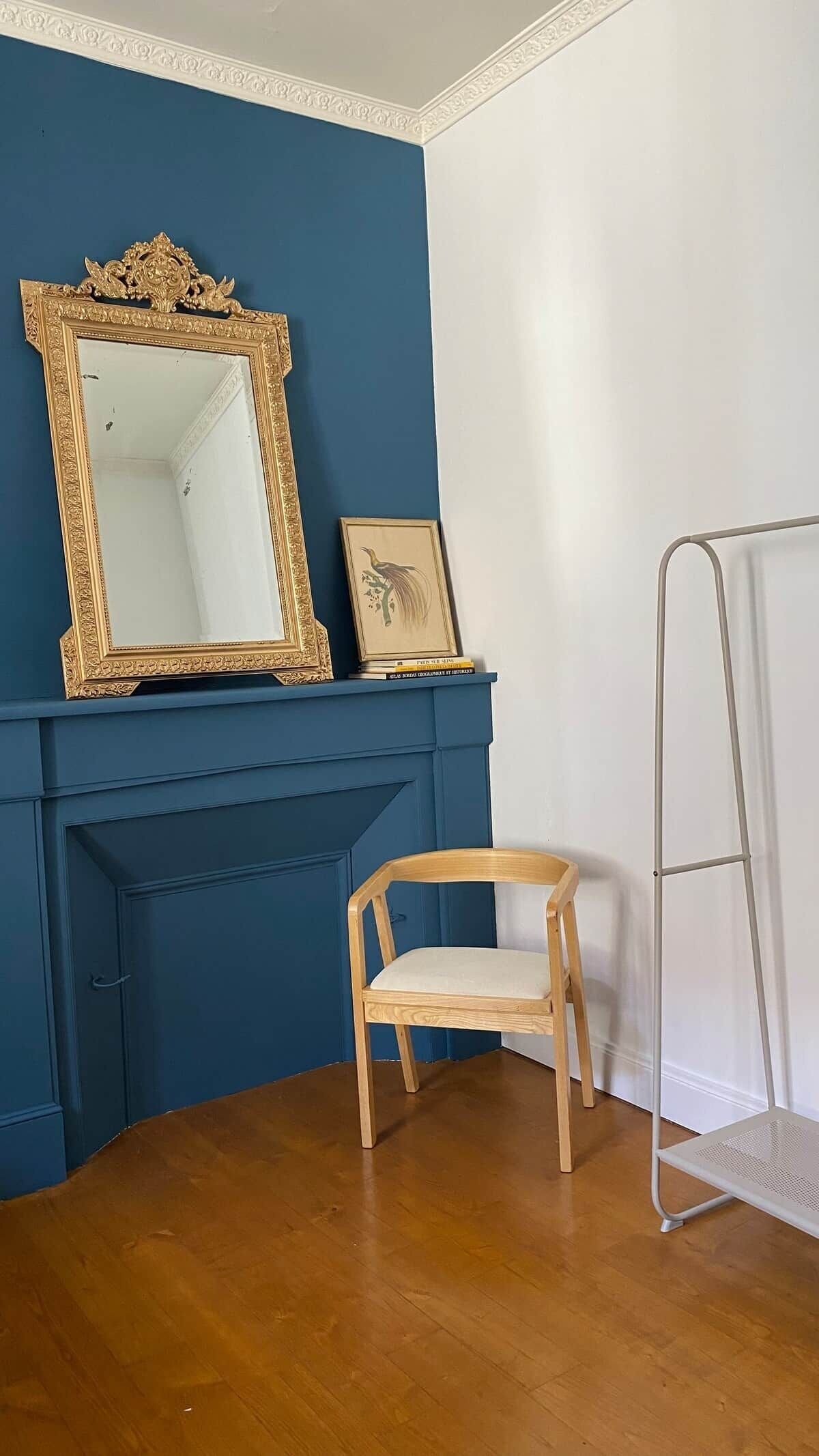 A room with a blue wall, a wooden chair, a metal ladder, and a fireplace with a gold-framed mirror and a framed picture.