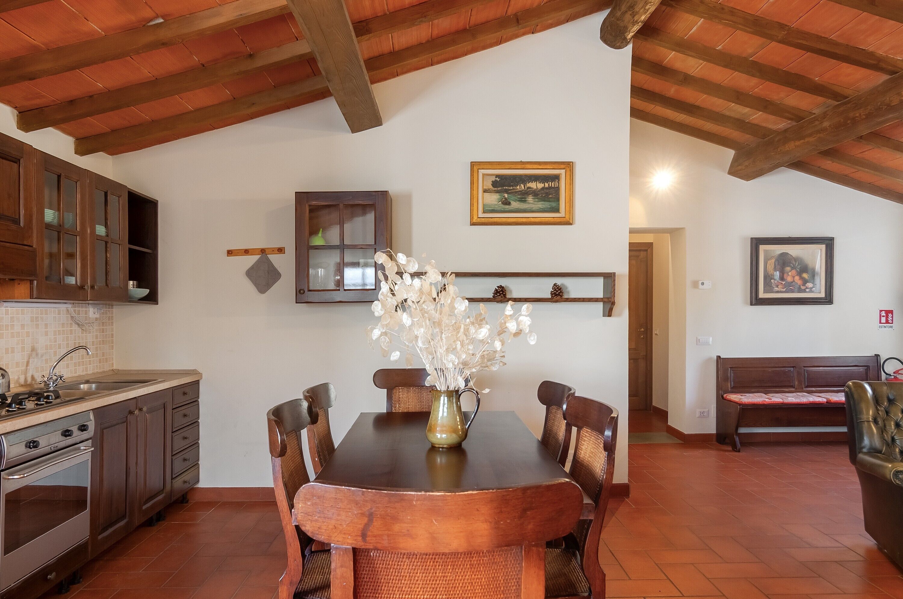 A kitchen with wooden cabinets, a dining table with chairs, a vase with flowers, and a painting on the wall.