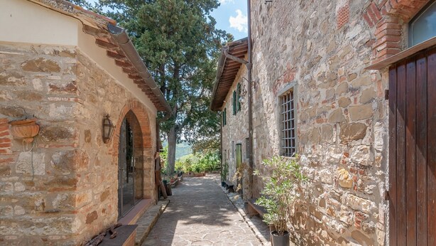 Un estrecho sendero de piedra entre edificios con puertas y ventanas de madera, que conduce a vistas de árboles y colinas.