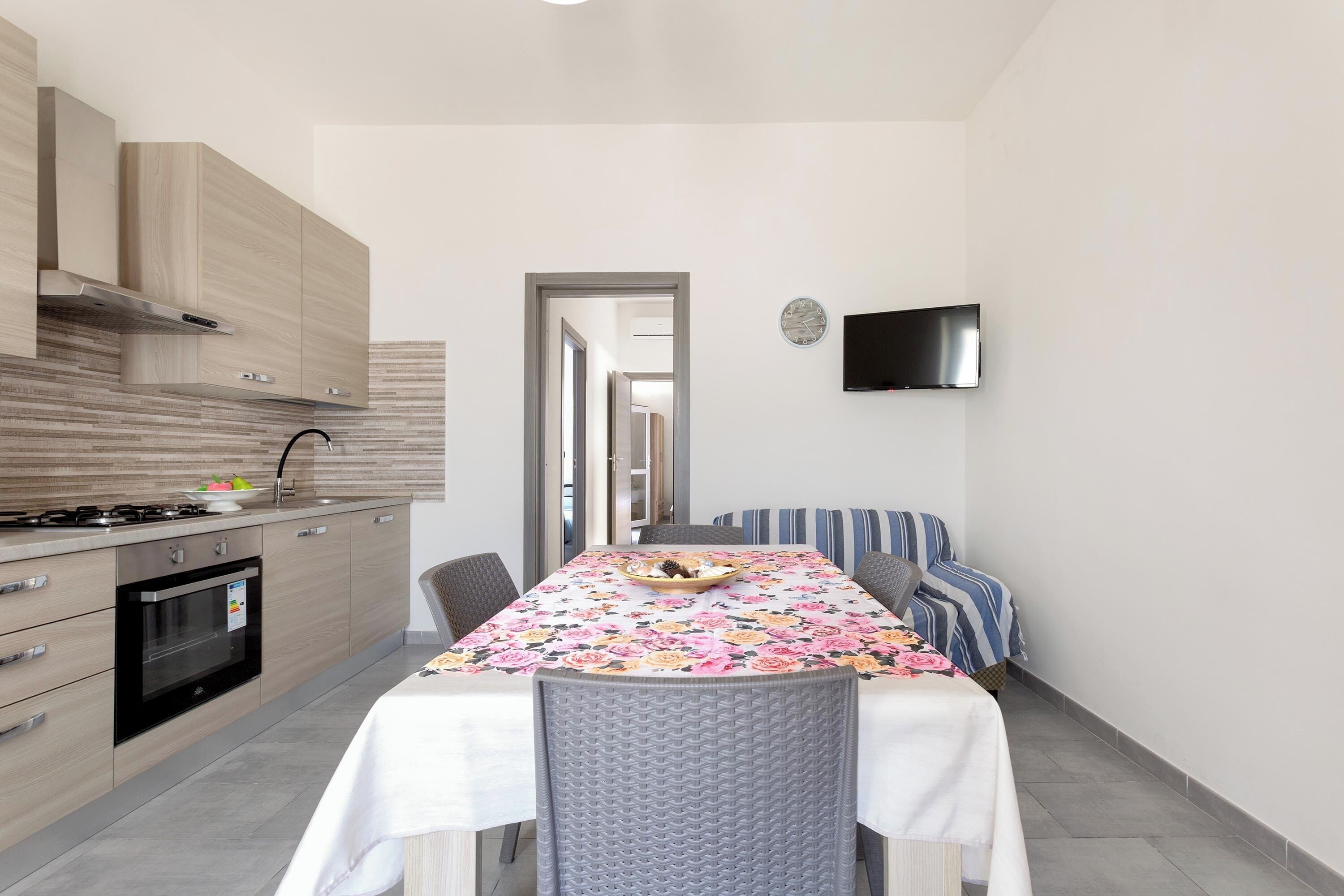 A modern kitchen with a dining area, a TV mounted on the wall, and a view into the adjacent room.