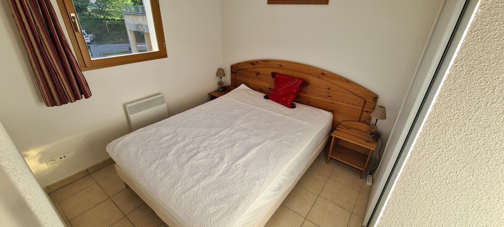 Una camera da letto con un letto, una testiera in legno, un comodino, un cuscino rosso, una finestra e un termosifone.