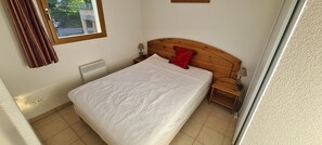 Un dormitorio con una cama, cabecera de madera, mesita de noche, almohada roja, ventana y radiador.