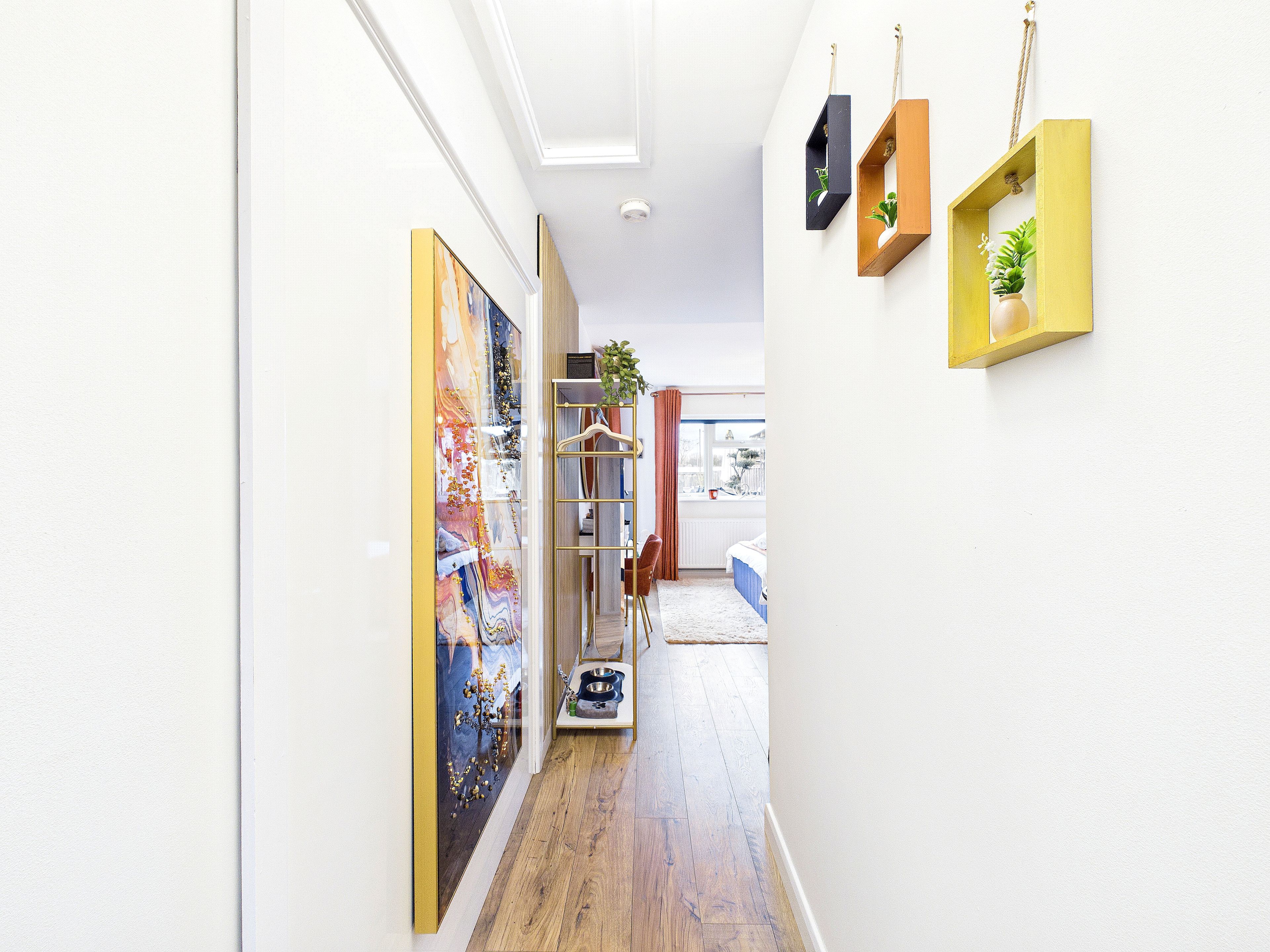 A hallway with a wooden floor, a bed with a blue bedspread, a window with a view, and a door leading to another room.