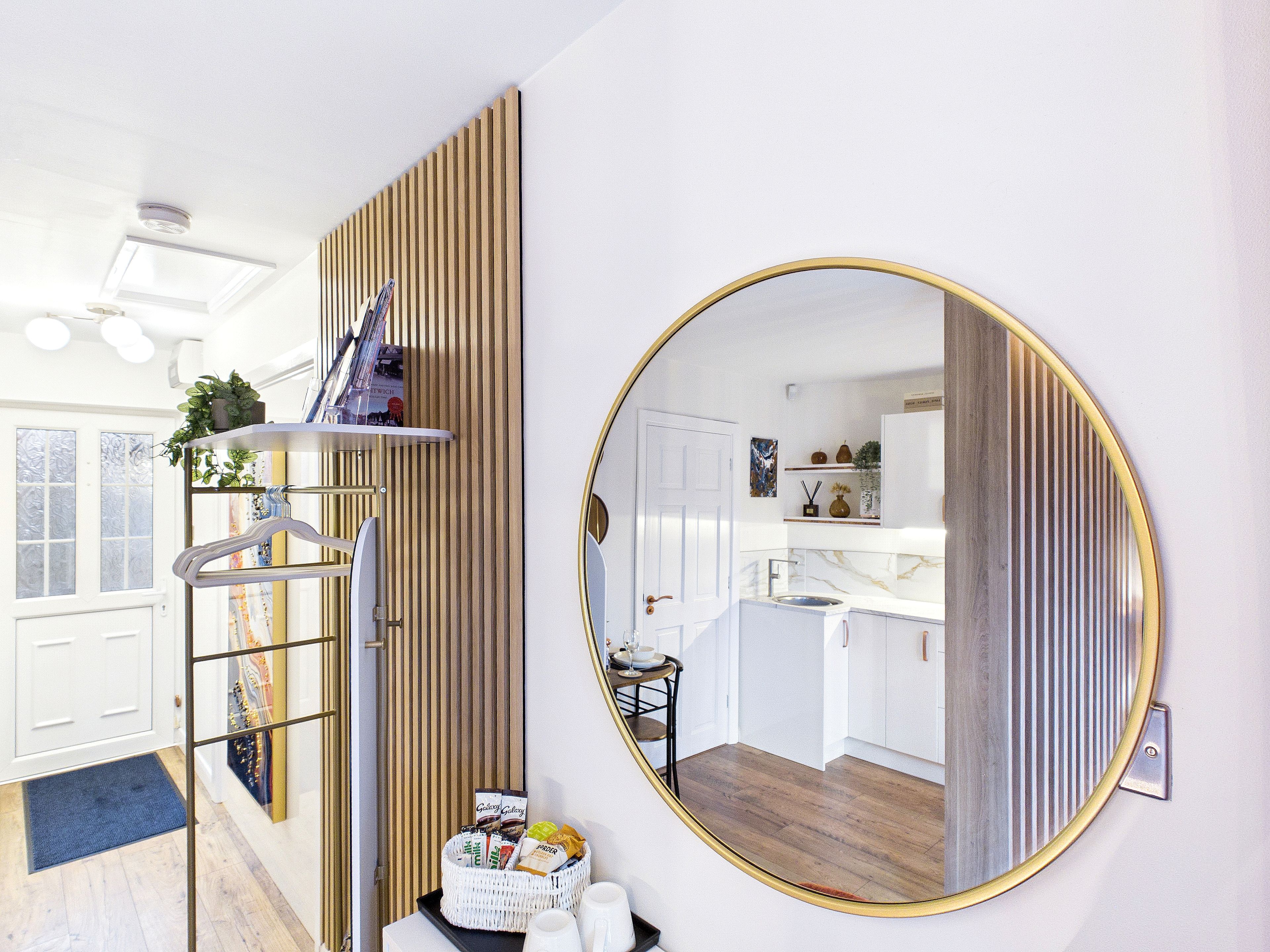 Une cuisine moderne avec des meubles blancs, un mur lambrissé en bois et un grand miroir rond doté d’un cadre doré.