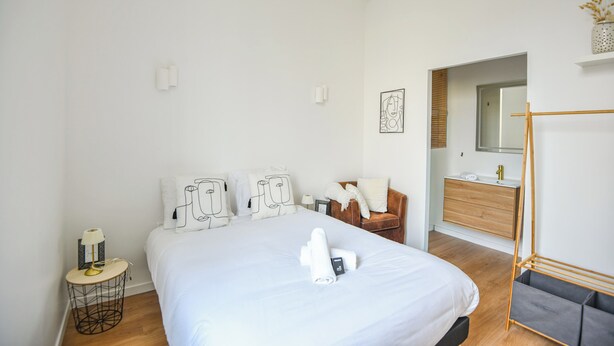 A modern hotel room with a large bed, a wooden dresser, a small round table with a lamp, and a bathroom visible through an open door. - Coeur Endoume - Bel appart 1 ch proche Vieux Port (Marseille)