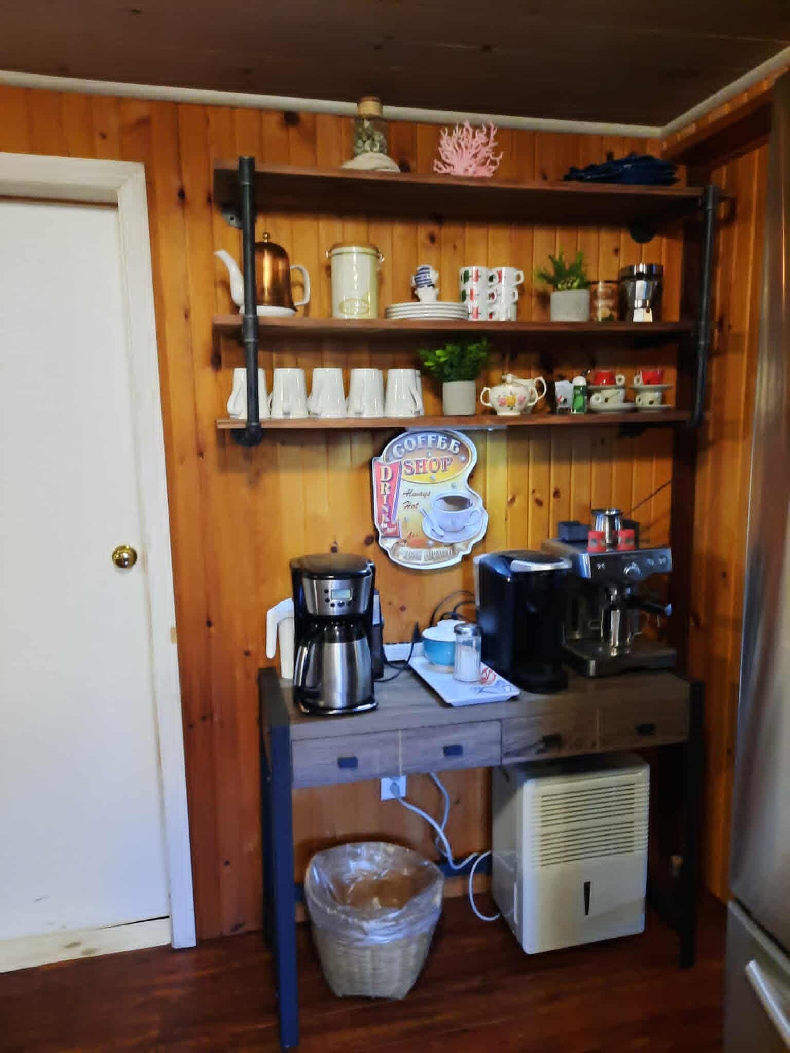 Cocina compacta con paredes de madera, un letrero de cafetería, una cafetera, un hervidor y un refrigerador pequeño.
