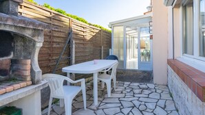 A patio with a stone floor, white table, and chairs, a brick fireplace, and a wooden fence with greenery. - Holiday Home “Quartier Calme” with Private Garden (Royan)