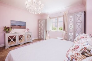 Ein Schlafzimmer mit einem großen Bett, einem Schminktisch, einer Chandelierleuchtre, einem Fenster mit Vorhängen und einem Gemälde an der Wand.