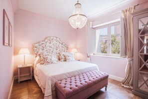 Een slaapkamer met een bed, nachtlampjes, een kroonluchter, een raam met gordijnen en uitzicht op het omliggende landschap.