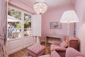 Ein zartrosa eingerichtetes Zimmer mit großem Fenster, einer Kristallleuchtre, einem Schreibtisch und einem Sofa.