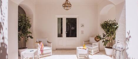 Una habitación con paredes blancas, una puerta, dos sillas de mimbre, una mesita y plantas en macetas.